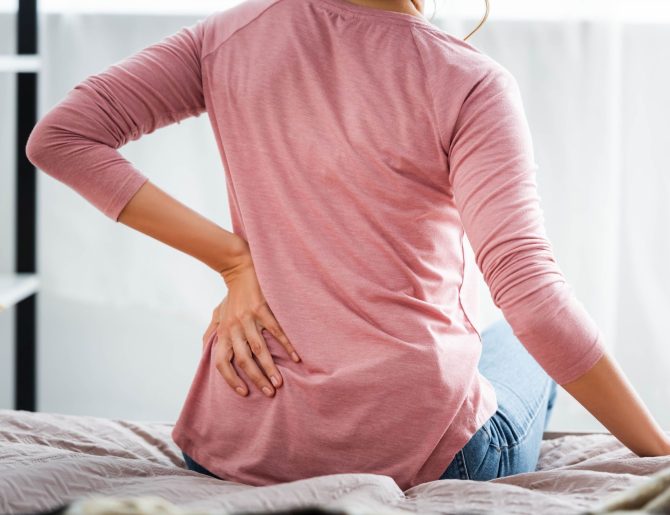 back view of woman with pain in back sitting on bed in apartment